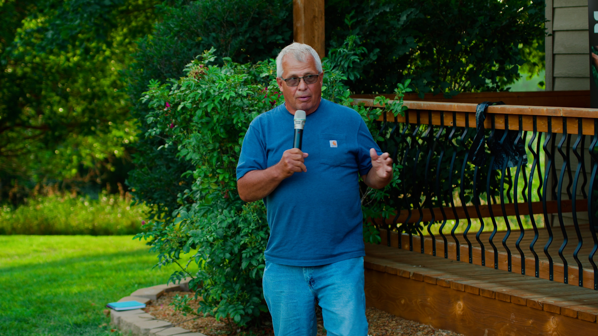 Dan Forgey speaks in front of a front porch while holding a microphone.