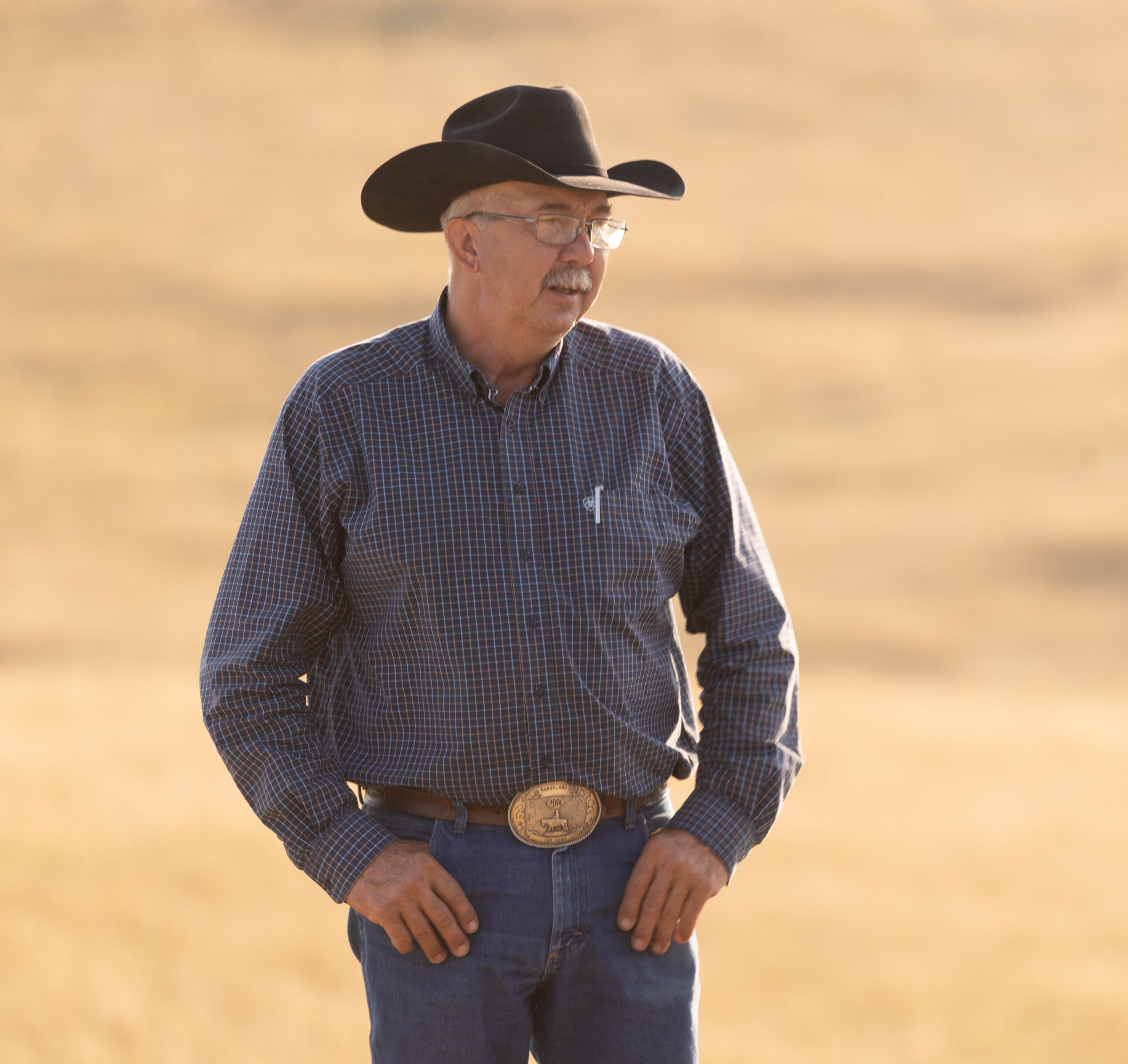 Rancher Ollila wears a cowboy hat and stands in a dormant pasture with his thumbs tucked into his blue jean pockets.