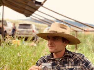 A photo of Cooper Hibbard seated tall grass wearing a cowboy hat and holding a glass mug.
